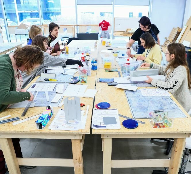 Kilka osób pracuje przy dużym stole warsztatowym, wycinając i składając elementy z papieru oraz innych materiałów.