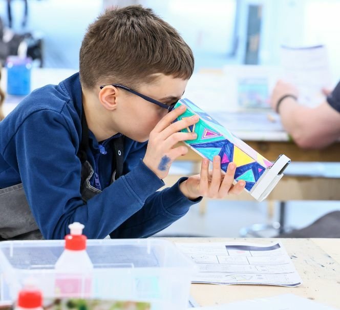 Chłopiec w okularach patrzy przez kolorowy kalejdoskop wykonany podczas warsztatów przy stole z materiałami plastycznymi.