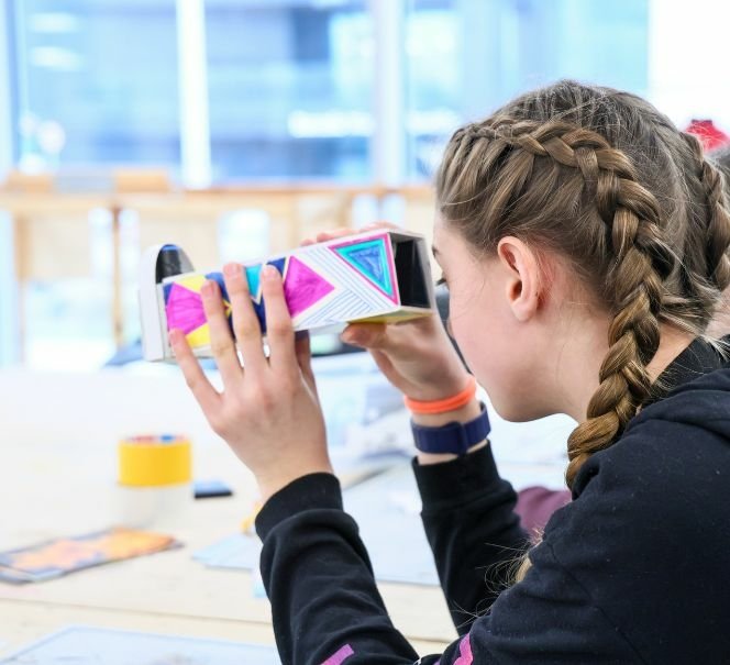 Dziewczynka z warkoczami patrzy przez kolorowy kalejdoskop wykonany z papieru podczas warsztatów przy stole z materiałami plastycznymi.