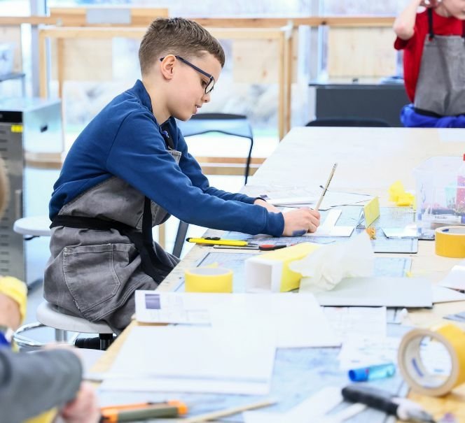 Chłopiec w fartuchu warsztatowym siedzi przy stole i rysuje elementy projektu na kartce, wokół leżą narzędzia i materiały do pracy.