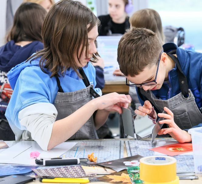 Dwoje dzieci w fartuchach warsztatowych wspólnie pracuje przy stole, składając elementy z kartonu i papieru.
