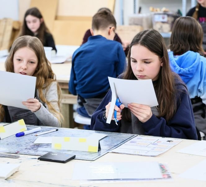 Dwie dziewczynki siedzą przy stole warsztatowym i wycinają kształty z papieru, przygotowując elementy pracy plastycznej.