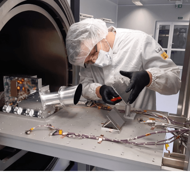 Naukowiec w stroju laboratoryjnym montuje element aparatury badawczej z przewodami przy dużym urządzeniu przypominającym tomograf.