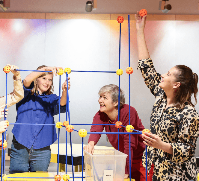 Trzy osoby budują przestrzenną konstrukcję z kolorowych łączników i prętów, wspólnie eksperymentując i dopasowując elementy.