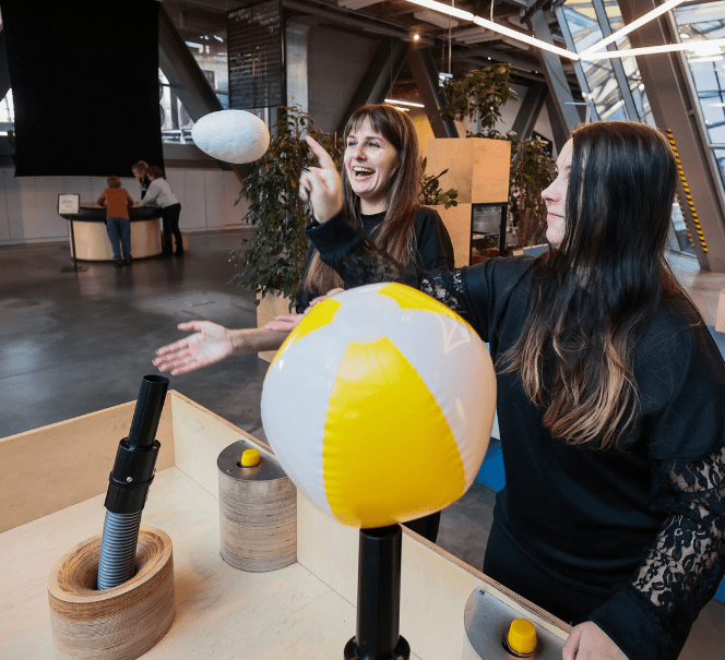 Dwie kobiety eksperymentują przy stanowisku z nadmuchem powietrza, unosząc lekkie przedmioty i piłkę w przestrzeni.
