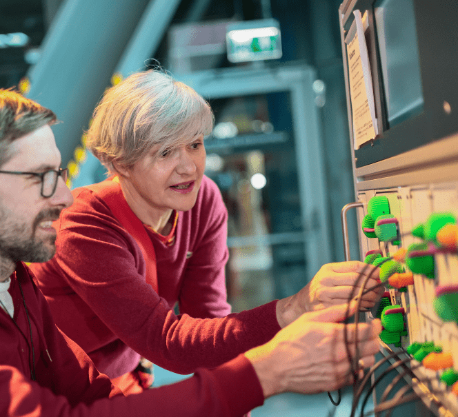 Kobieta i mężczyzna manipulują elementami instalacji z kolorowymi pokrętłami i przewodami, testując działanie urządzenia.