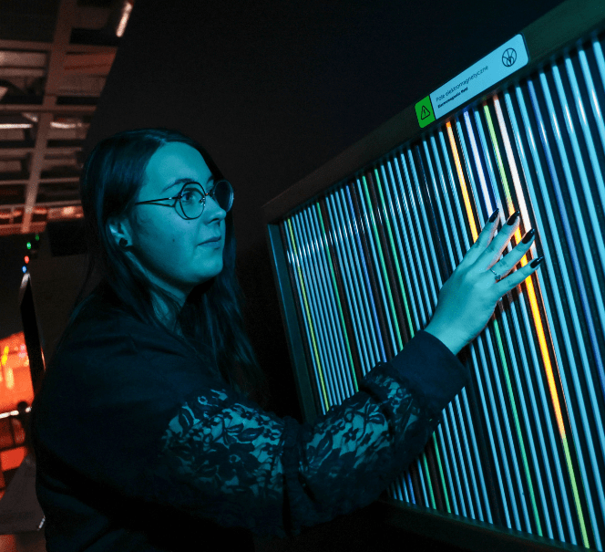 Kobieta dotyka podświetlonej ściany z pionowymi liniami, zmieniając ich kolor i układ światła.