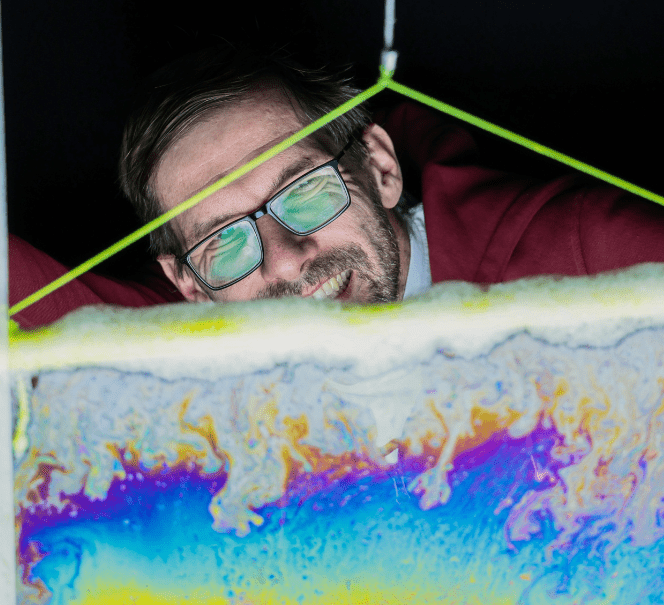 Mężczyzna obserwuje cienką warstwę cieczy o tęczowych barwach, tworzącą abstrakcyjne wzory.
