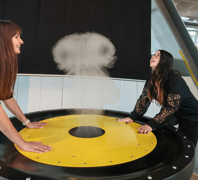 Dwie kobiety stoją przy okrągłym stanowisku, nad którym unosi się para wodna, tworząc kształt przypominający chmurę.
