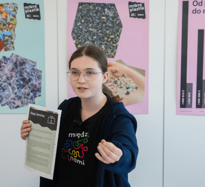 Uczennica prezentuje wyniki projektu o mikroplastiku, trzyma kartę pracy; w tle plansze edukacyjne i wykresy.