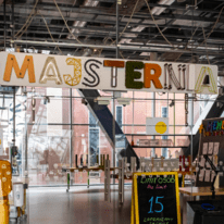 Zdjęcie wejścia do Majsterni, napis jest złożony z różnych elementów jak deski, światła led, mech...