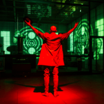 Ciemne pomieszczenie, na środku stoi tyłem podświetlona na czerwono osoba z uniesionymi rękami.